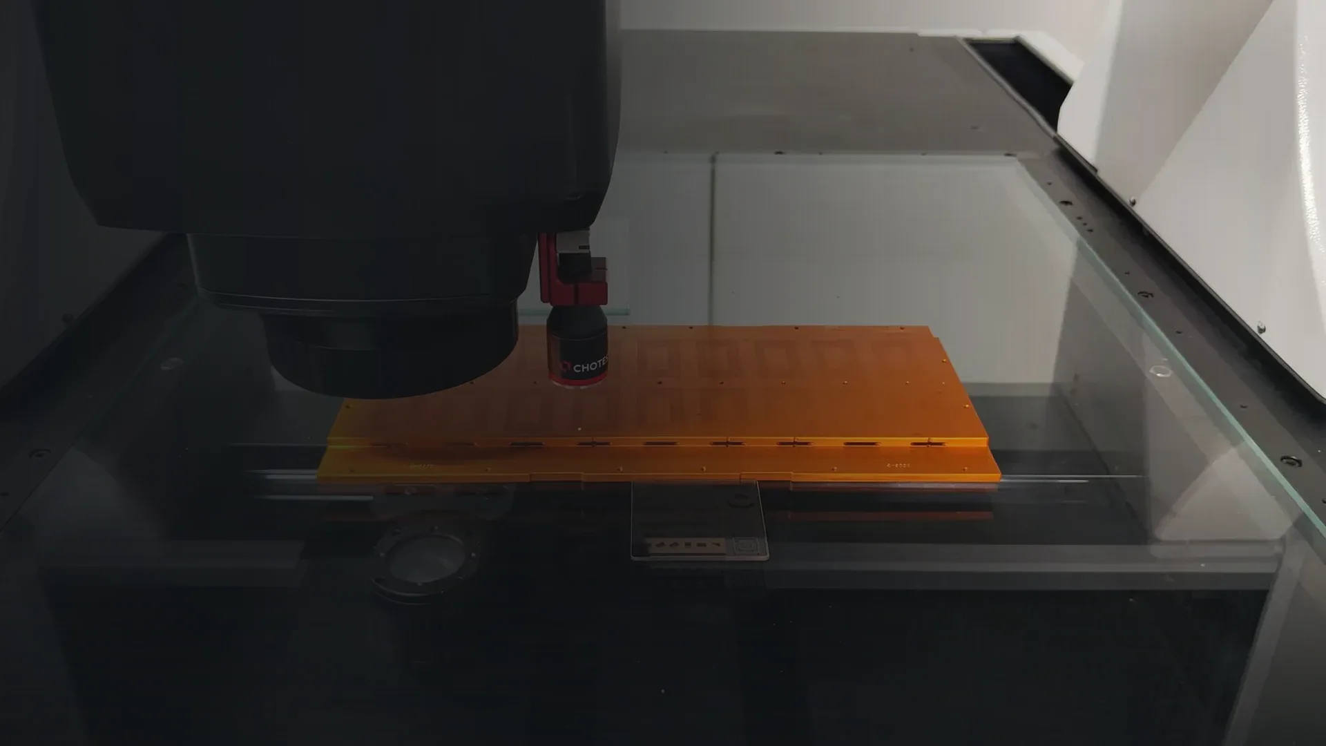 Messkopf eines Chotest-Novator-Bildverarbeitungsmessgeräts über einem orangefarbenen Werkstück auf einer Glasauflage